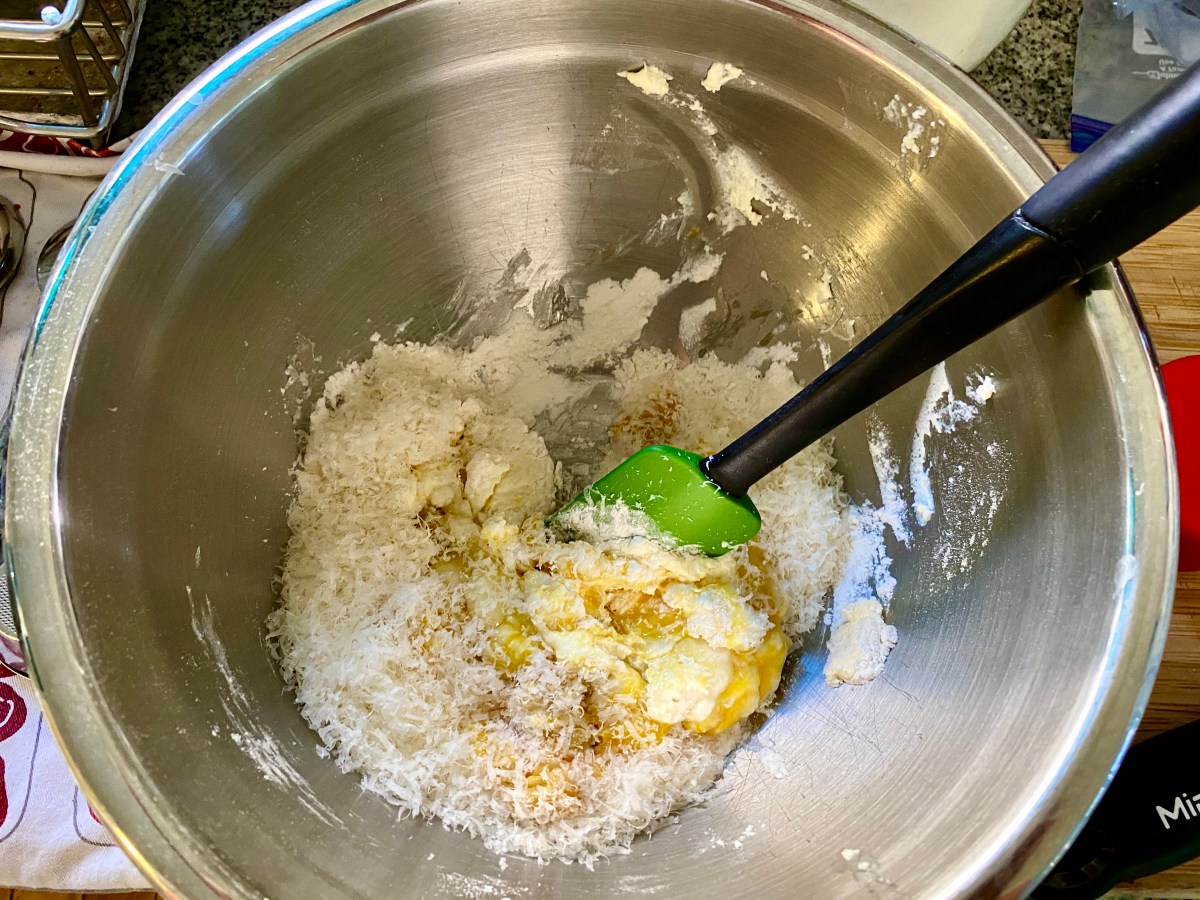 A silver bowl with a messy shaggy mix of flour, eggs, ricotta cheese, and salt just starting to get stirred up with a green silicon spatula