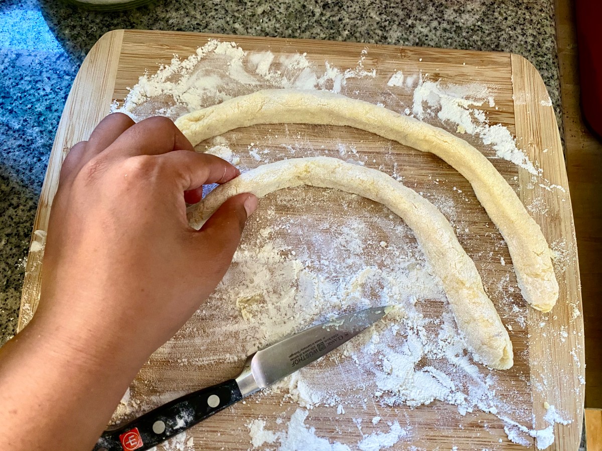 On a flour board, a brown hand pinches at the long arced snake of gnocchi dough that has just been rolled out