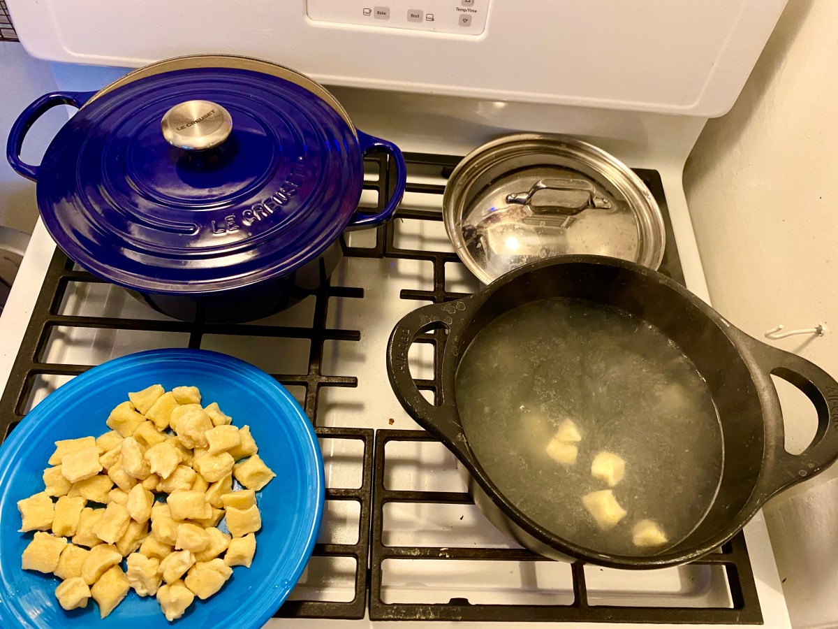 An overhead shot of the stove top. On the top left is a blue Le Creuset, where the short ribs are simmering. On the burner in front of that is a light blue plate where cooked gnocchi are piled high. And to the right of the plate is where a boiling pot of water is cooking raw a few pieces of gnocchi. You can see them started to emerge from the cloudy water.