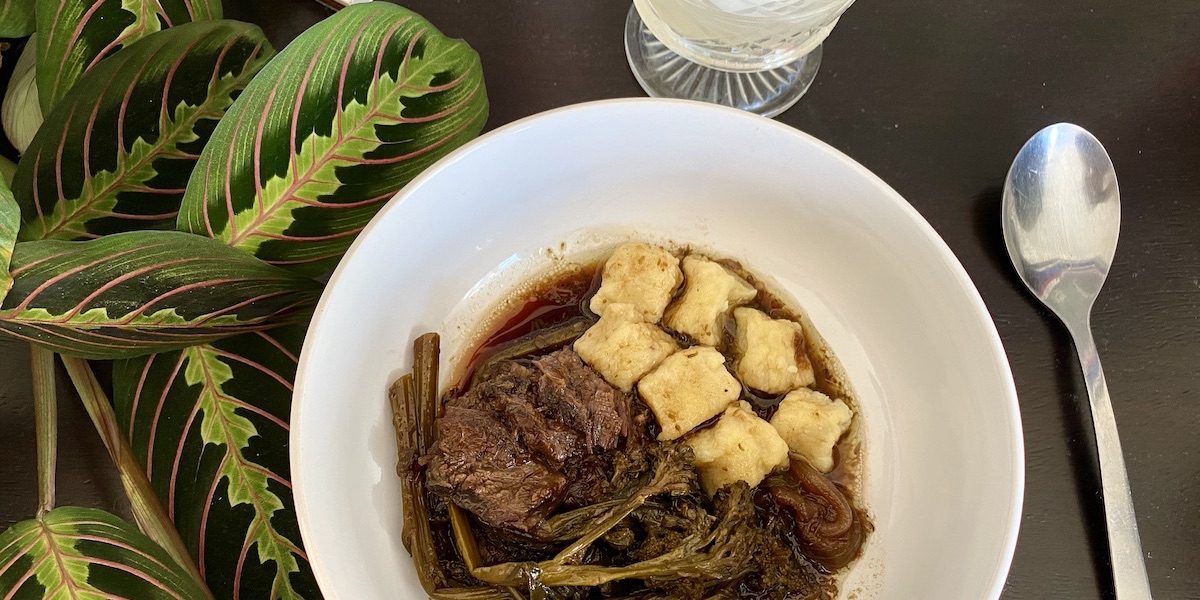 A bowl of braised beef, broccolini, and ricotta gnocchi sits on a black surface, and the leaves of a prayer plant snuggle up against it. A spoon sits next to the bowl and a cut crystal glass peeks into the edge of the frame. It's time to eat.
