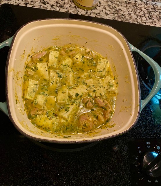 a square dutch oven filled with cubes of paneer and chicken, a yellow marinade and flecks of green herbs cooking on the stove