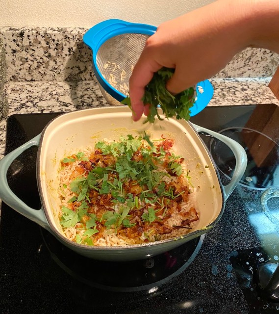 a hand sprinkles herbs over the top of a dutch oven that has been filled with rice and sprinkled with fried onions