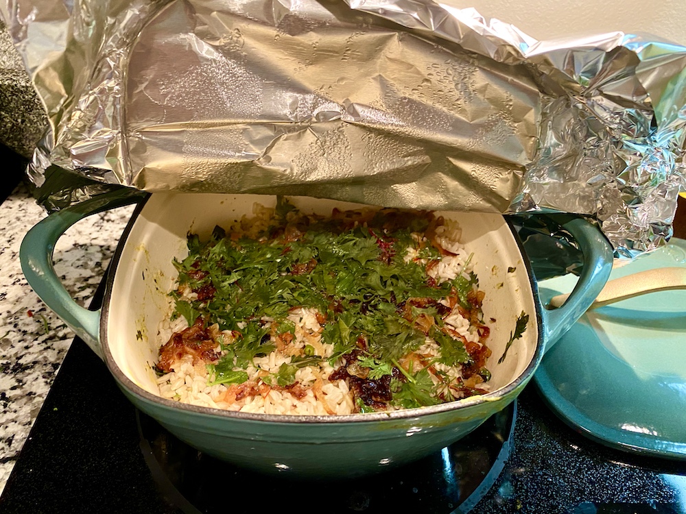 a foil cover is lifted open on a dutch oven, revealing a smattering of dark fried onions and herbs over rice: the top of a biriyani! 