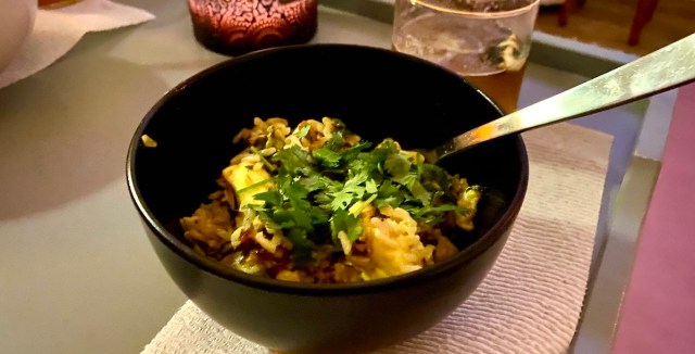 a black bowl holds a serving of biryani. yellow rice and green herbs stand out from the bowl, and a soft purplish candle glows in the background behind it