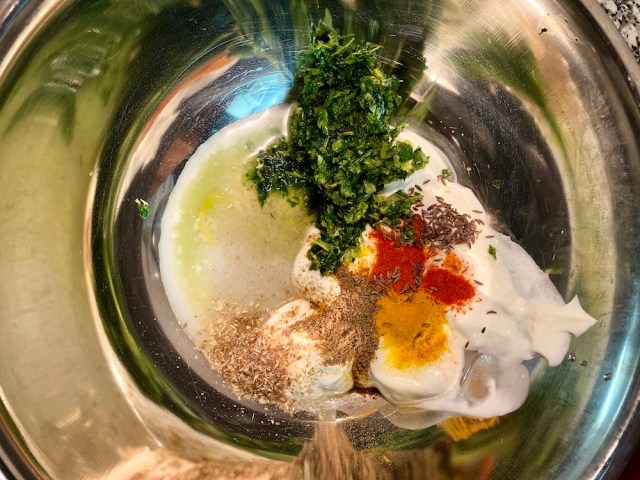 a silver bowl with a plop of white yogurt inside, the green paste from the mortar, a yellow pop of turmeric, a red flash of chili powder and some other browner patches of spices, about to get mixed together for the marinade