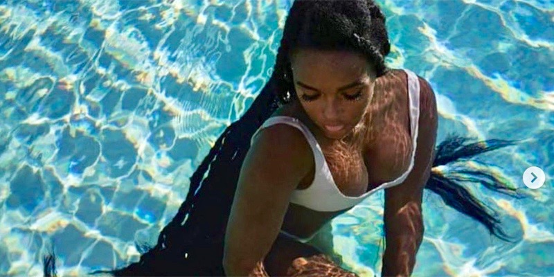 Janelle Monae lounges in water in a small bathing suit. Her hair floats around her in the aqua marine water that dances against her skin.