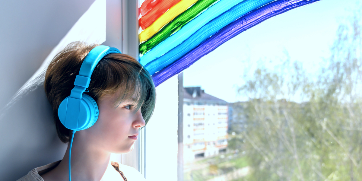 A sad teenage lesbian full of loneliness and missing her friendships, listening to a playlist on her iPhone while looking out a window with a handprinted rainbow on it.