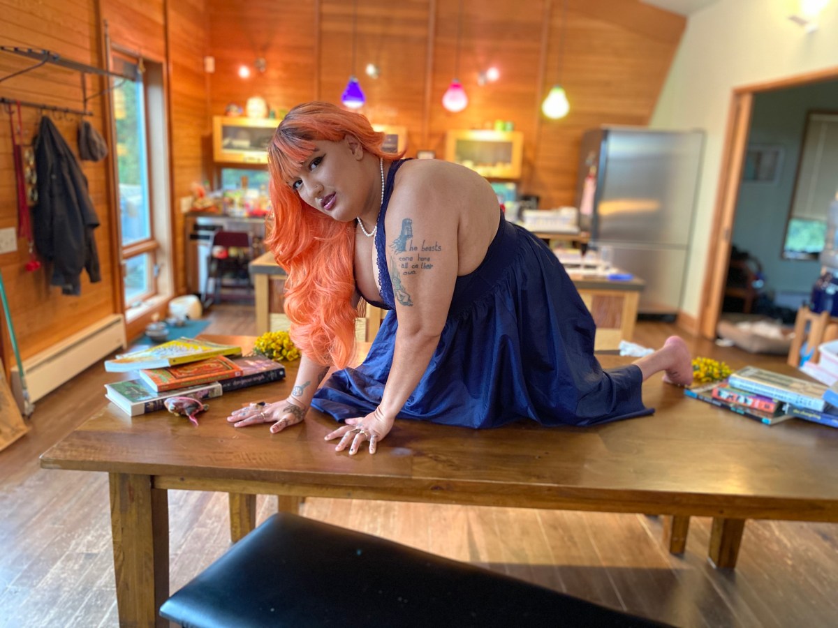 Neve is a light skinned person with pink hair and is posing on a table.