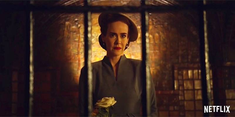 Sarah Paulson as Nurse Ratched stares horrifically behind bars in captivity, with bricks behind her glowing yellow.