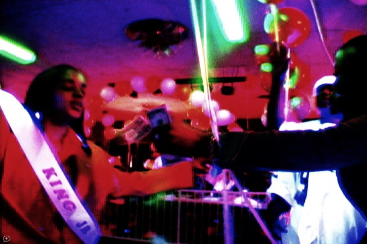 a person in a pageant sash, which reads "King Jr." takes a small stack of bills from a hand that's reaching into the screen.