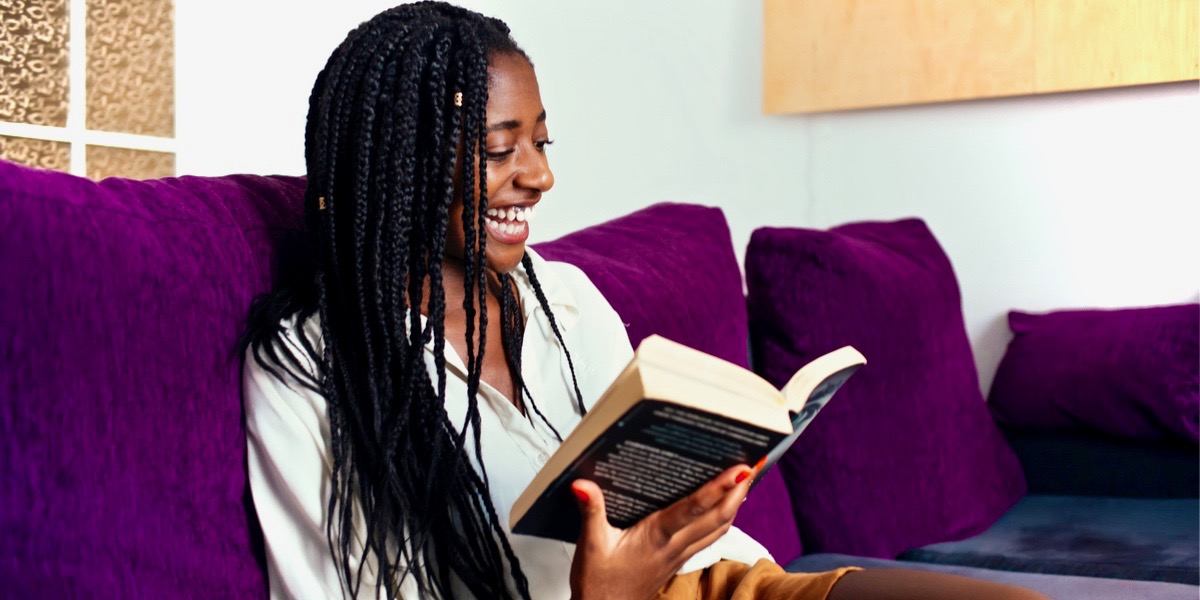A cheerful young black woman with long braided hair is reading a thick book on a purple couch.