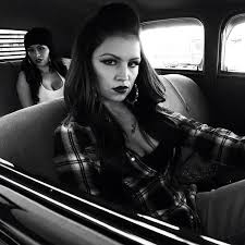 a chola with sharp eye liner, eyebrows, lipstick and big hoops, sits in a car with her friend in the back and glowers out of the passenger window