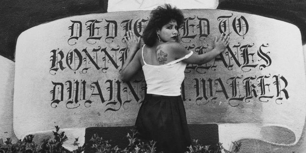 A tough and pretty chola woman, with the top of her hair teased and dark lipstick, places both of her hands on a huge painted mural banner and looks over her shoulder with anguish and "don't fuck with me" energy. She is looking over her shoulder and the top of her white tank top is pulled to the side showing off a tattoo. The banner, in a stylized script reads: "Dedicated to Ronnie ..anes Dwayne Walker."