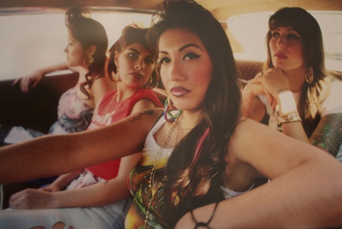 four chola women ride inside of a car together. the one in the driver's seat has a hand on the wheel, an elbow perched on the window sill and a made up face with winged eyeliner and dark lipliner