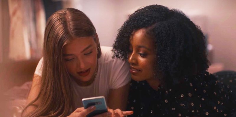 Olivia and Amber look at a phone together while lying on the same bed