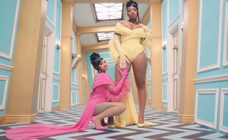 In the hallway of a mansion, Cardi B wears a pink bodysuit with a train, and kneels below Megan The Stallion, who stands prominently in the center, in a matching yellow bodysuit, looking like a boss. They both have gloved hands that meet on Meg's thigh, and are taunting the camera with sexily menacing stares..
