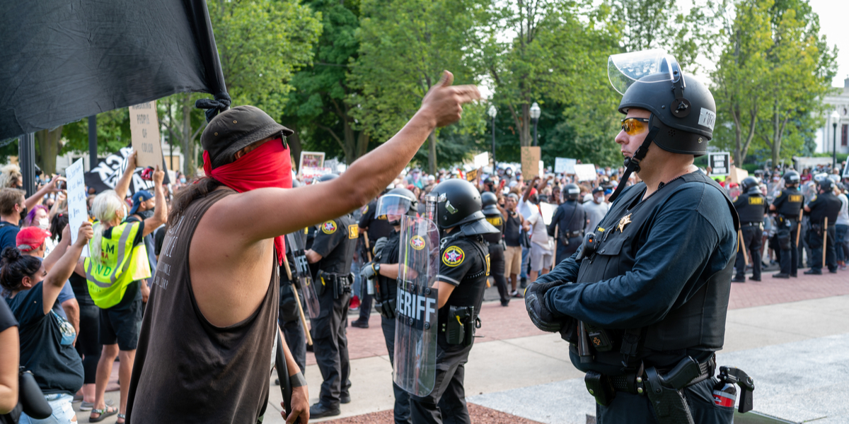 A standoff in Kenosha between protestors on the left and police in riot gear on the right; in the foreground, a white person wearing a red bandana as a mask gestures angrily at a cop, who looks on impassively
