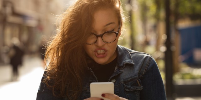 A frustrated-looking white woman wearing glasses and scowls down at her phone as she types.