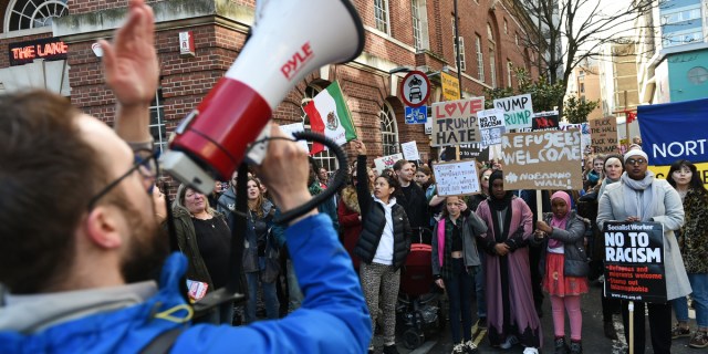 A person with a megaphone in the foreground addresses a group of protesters holding signs opposing the Trump administration's anti-LGBTQ policies.