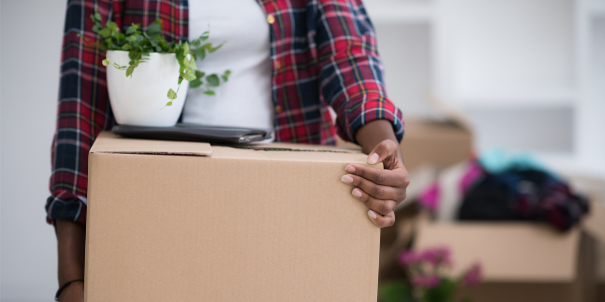 A Black woman in flannel holding a box with a small plant on top of it. Half-packed boxes are in the background because she is in the process of moving.
