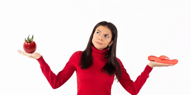 A woman in a red turtle neck has her arms outstretched, one holding a red tomato and the other holding a red rabbit style vibrator.