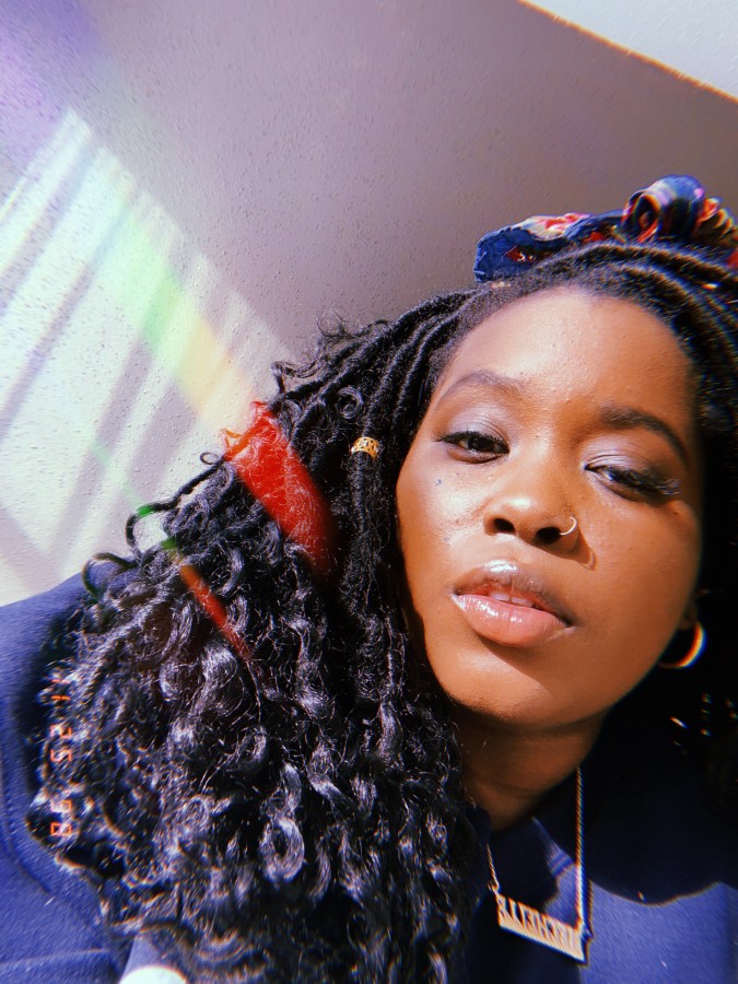 A selfie of Shelli looking at the camera with lips parted, wearing a scarf wrapped around her head over faux locs and a nameplate necklace with her lips parted; the photo is dreamily edited with the HUJI app.