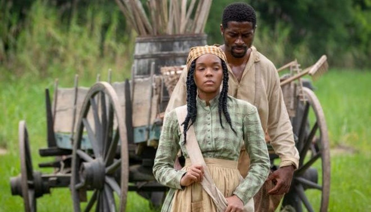 Janelle Monáe as Eden in "Antebellum." Eden walks on the plantation where she lives. Her braided hair is tied back with a head wrap and she wears a green checkered dress with a khaki apron.