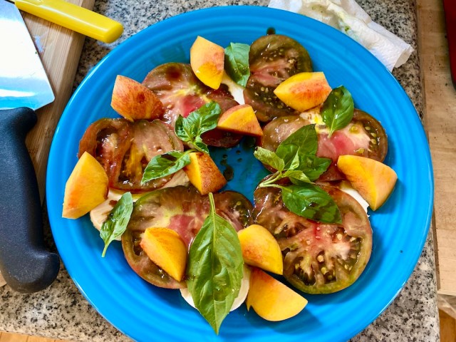 a bright, light blue plate with a circle of red tomatoes, orange nectarines, green basil leaves and peeks of white mozzarella underneath