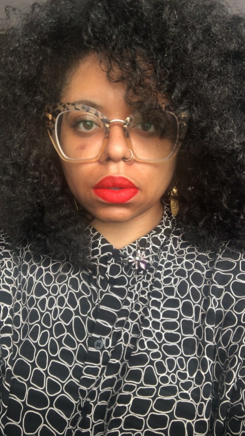 A closeup selfie of Dani looking directly into the camera wearing red lipstick and leopard print glasses frames; her hair fills the rest of the frame around her head, and she's wearing a black and white patterned high-neck top.