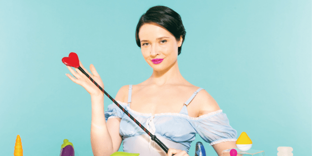 A woman in a blue dress stands in front of a table of sex toys and holds a heart-shaped riding crop.