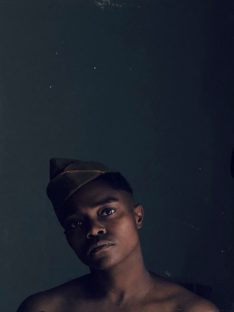 a photo of the author's head in peaked military cap, and bare Black shoulders. he stands in a darkened room with just a small shine of light playing off his cheekbones and jaw.