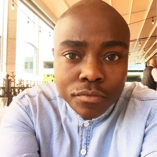 a photo of the author from the shoulders up, he's wearing a light blue button-up shirt, buttoned to the top. he is Black and bald with thick eyebrows, big brown eyes, and he looks into the camera with a slight bemusement and lift of the left side of the lip.