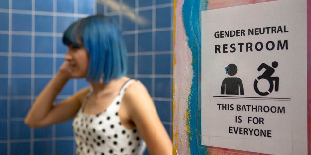 A nonbinary femme person with blue short hair stands in the background. In the foreground is a sign that reads, "gender neutral bathroom, this bathroom is for everyone."