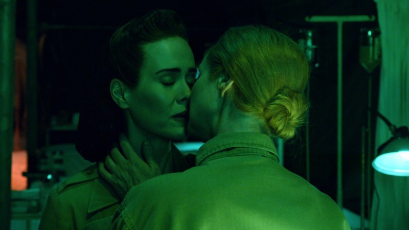 Sarah Paulson, as Nurse Mildred Ratched, leans into a romantic and steamy kiss with Cynthia Nixon as Gwendolyn Briggs in the Netflix series "Ratched." They're faces are awash in green lighting.