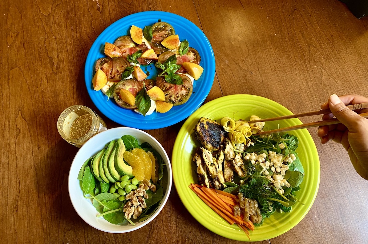 three salads on a table: one with avocado and orange in a small bowl in the bottom left ocrner, one on a bright blue plate with tomatoes , mozzarella and basil, and one on a bright green plate with chicken, carrots, corn kernals on top of a bed of greens