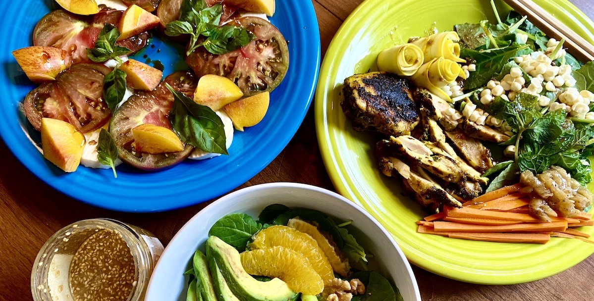 three dishes sit on a table touching their edges. a bowl with a peek of avocado and orange is at the bottom, a blue plate with tomatoes and nectarines is on the top right, and a bright green plate with chicken, corn, carrots and greens, and small rolls of yellow squash.