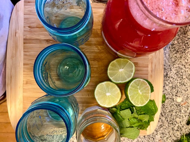 an overhead shot of the open mouths of three blue glass jars, three lime halves, a bunch of mint, a glass har of thick simple syrup, and a pink jar of watermelon juice