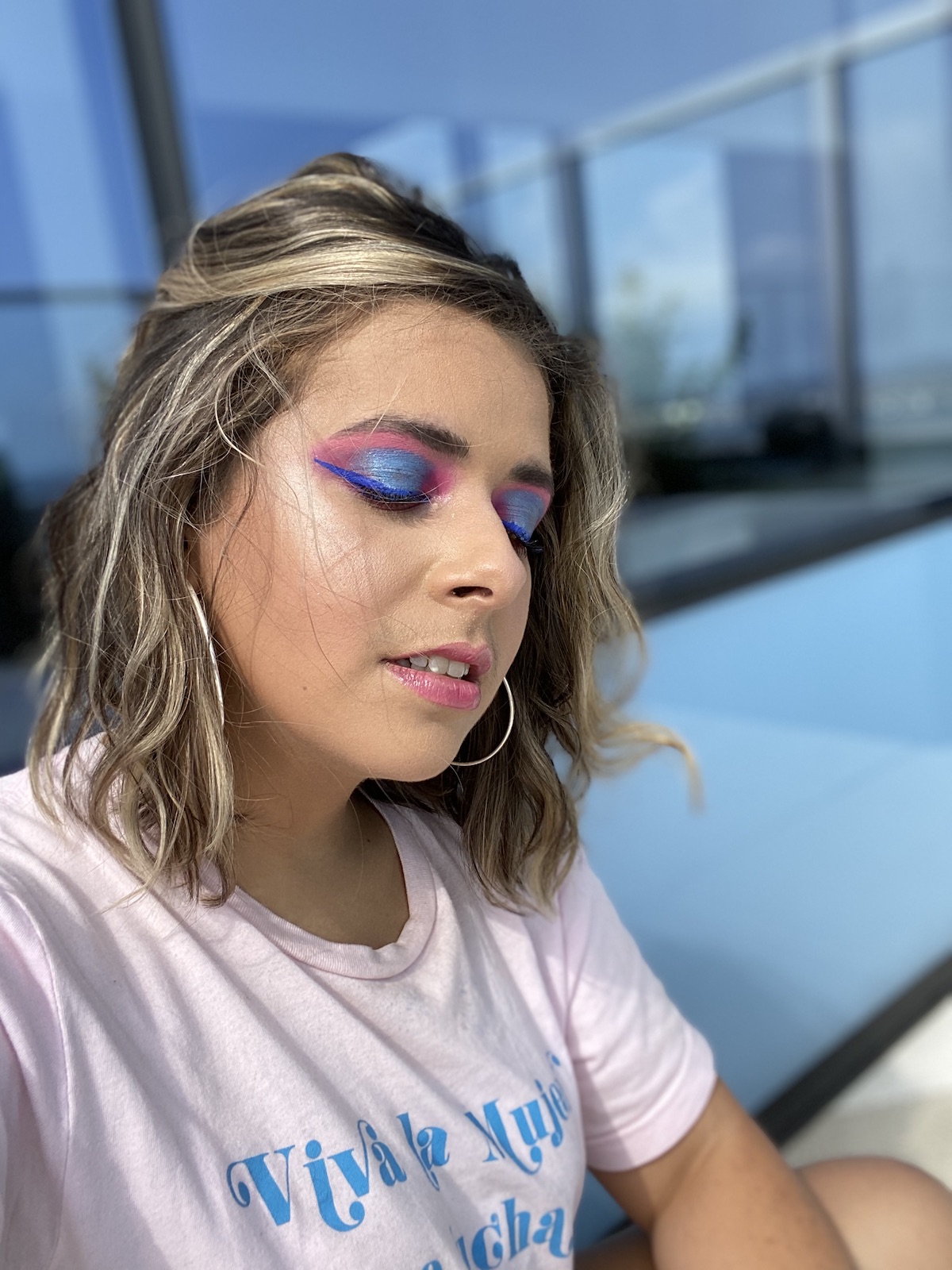 The author, Olivia Zayas Ryan, poses with a full face of makeup. Her eyes are closed and the makeup is dramatic, blue and pink. She wears large hoop earrings and looks at peace.