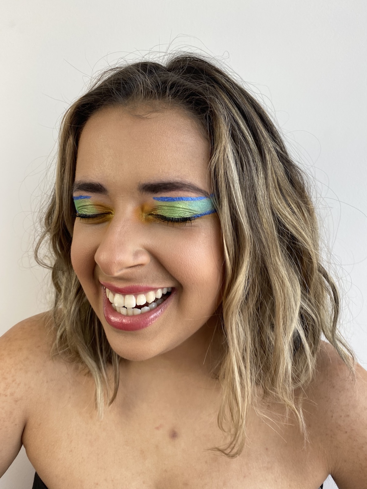The author, Olivia Zayas Ryan, is caught laughing and smiling with her mouth open, teeth showing. Her eyes are closed and she has dramatic makeup on, shades of green and blue and yellow. Her expression is happy.