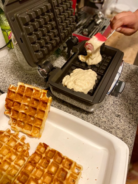 an open waffle iron and two clumpy batches of wet waffle dough on the bottom part of the press. in the foreground crispy cooked waffles stand in a tray