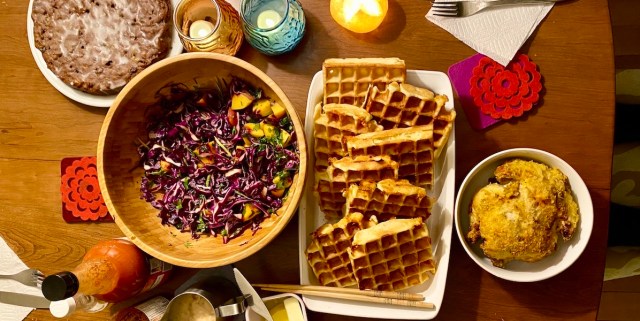 an overhead view of a table setting with a large bowl of purple cabbage salad, a tray of waffles, a pitcher of gravy, a small bowl with a little round fried hen nestled inside