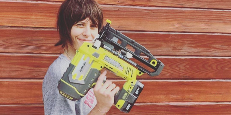 Kate Moennig stands in front of a wood panel wall with a very large power tool of some sort that's bigger than her own head!