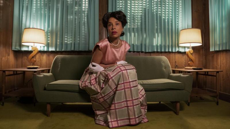 Sophie Okonedo as Charlotte in Netflix's "Ratched." She is in a shiny pink silk top and white gloves, along with a pink and grey plaid skirt in the same shiny fabric. She sits hunched over on a green couch in front of turquoise blue window curtains.