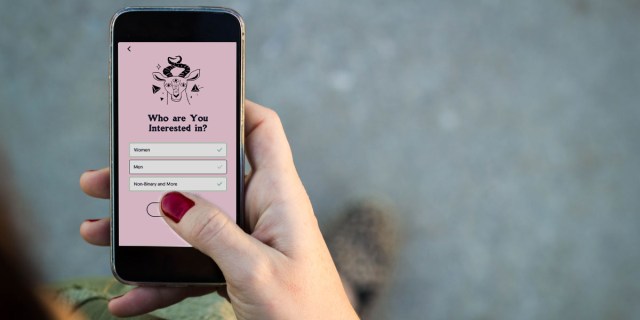 A hand of a white person with short, red-painted nails is holding a smartphone out in front of them; a whimsically illustrated pink menu that asks "Who are you interested in? Women/Men/Nonbinary and more"