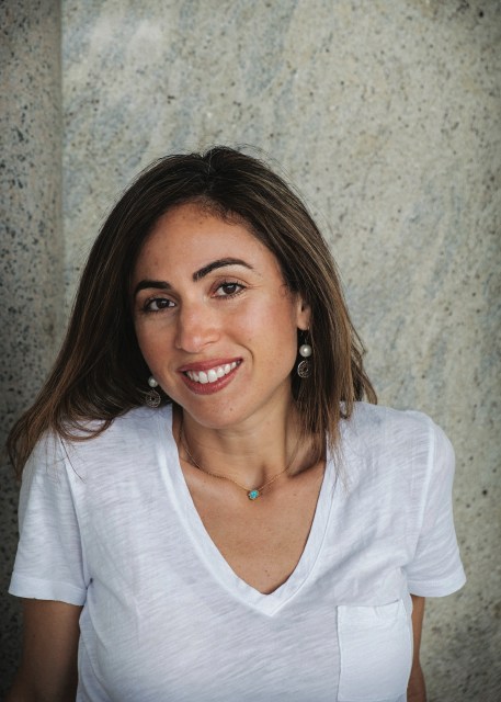 a Palestinian American woman in a white v-neck t-shirt stands in front of a wall with a half smile, her face turned just slightly away from the camera. she has light brown, straight shoulder length hair, dark eyebrows and a red lipstick smile