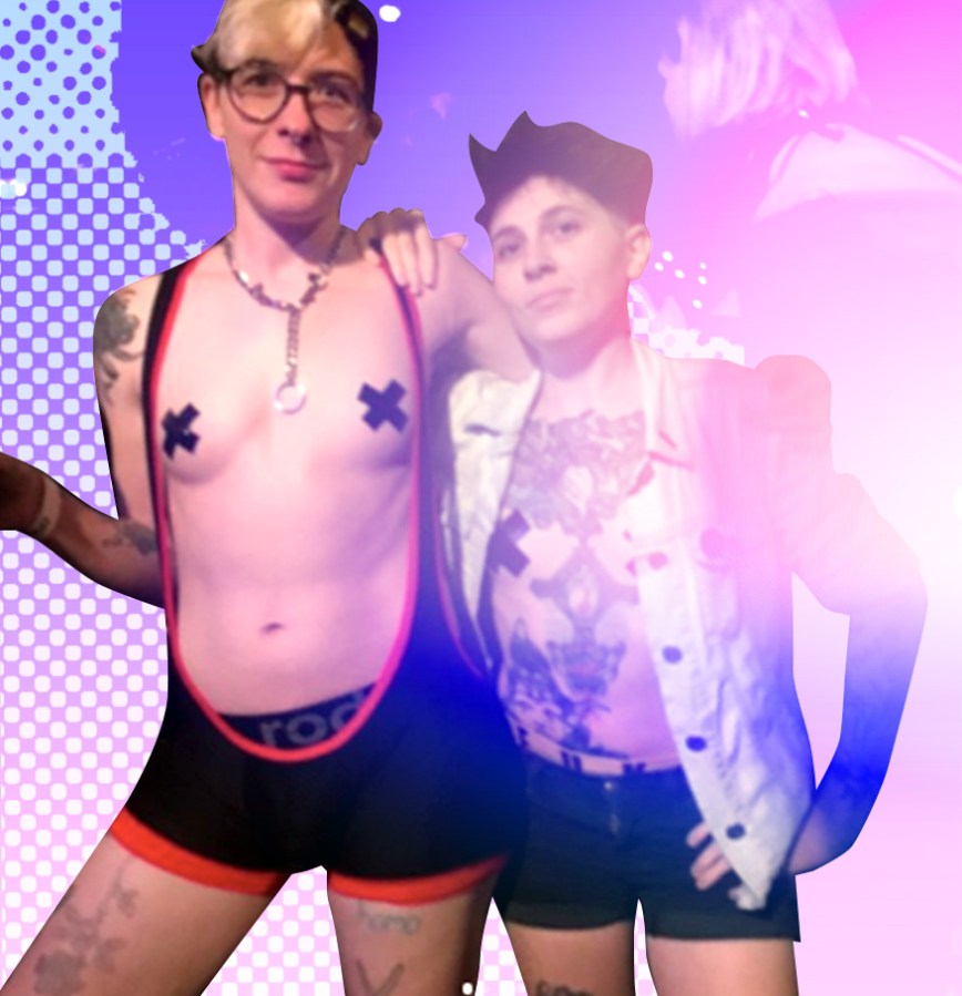 Archie poses with their partner, Gus, for the camera; Archie is wearing an open-front leotard with the hem of a Rodeoh harness showing beneath; their nipples are covered with black tape and a metal chain with an o-ring is around their neck. Gus is wearing black denim shorts and an open denim jacket, also with black tape covering their nipples and many tattoos visible.