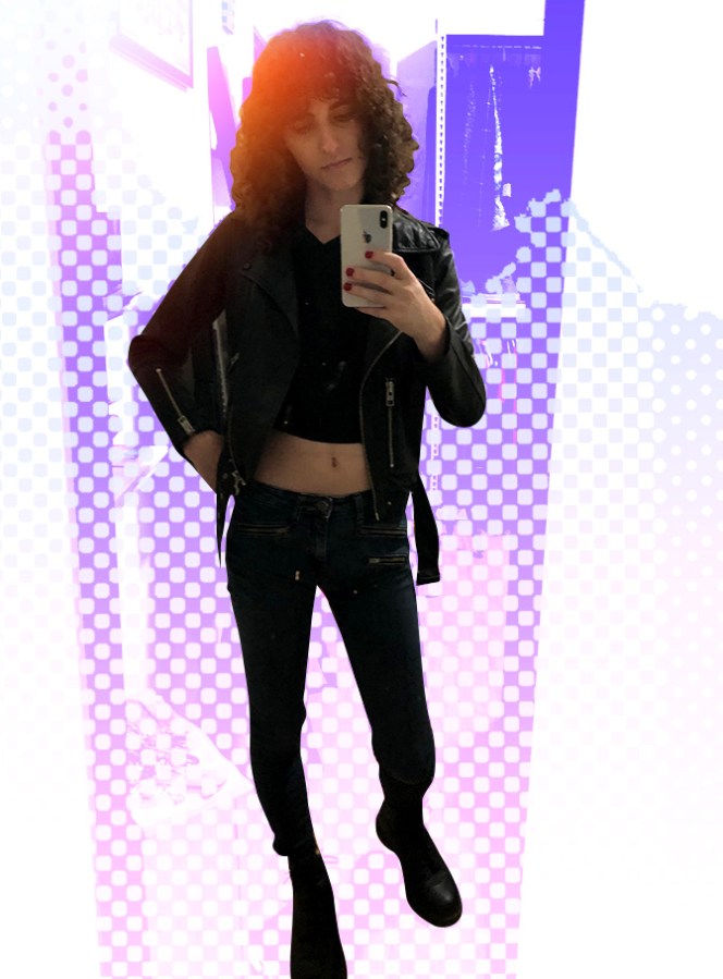A full-length mirror selfie of Drew, a thin white woman with shoulder-length dark curly hair, wearing a dark crop top, black leather jacket, black skinny jeans and boots