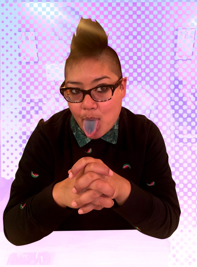 A photo of Kamala, a brown person wearing glasses with a mohawk, seated from the waist up. She's wearing a dark sweatshirt with a watermelon print pattern and a green patterned collared shirt peeking out, and has her hands clasped in front of her as she sticks out her tongue, which is stained blue.