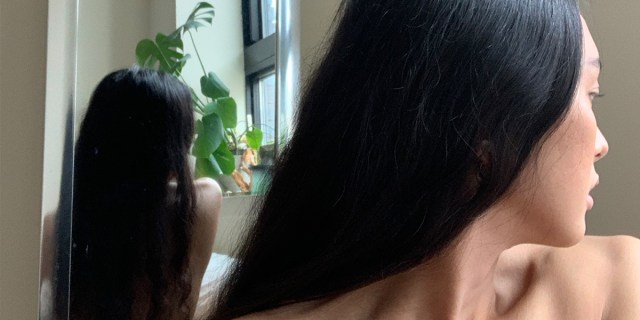 xoai pham looks over her bare shoulder, her image reflected in the mirror behind her. natural light at the windowsill filters over a leafy green houseplant.