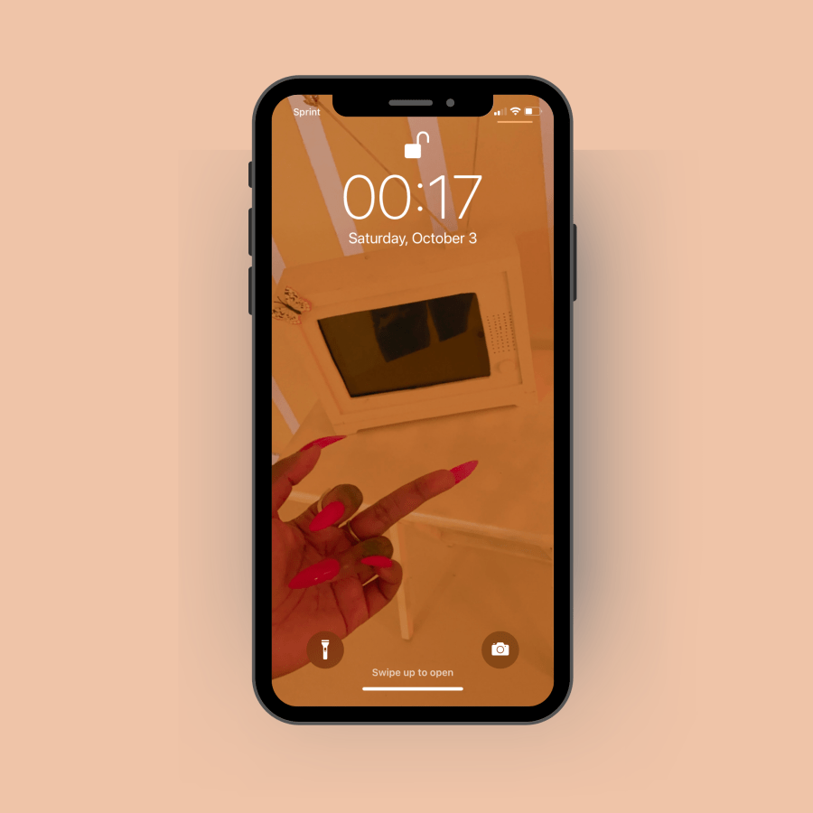 A screenshot of an iPhone lock screen. The photo is of a Black woman's hand with impeccable long red nails, giving the middle finger. It's set against a pastel Orange background.